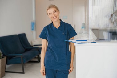 Klinikteki resepsiyonda elinde dosya olan bir doktor var ve kameraya bakıyor. Yüksek kalite fotoğraf