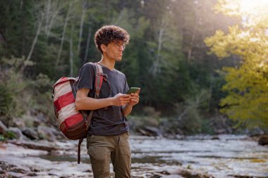 Man traveler with backpack looking for route on smartphone at river in mountains. Tourist in nature.