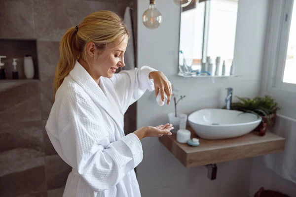 Young beautiful Caucasian woman pouring body lotion on hand in bathroom. Anti-aging concept. At home