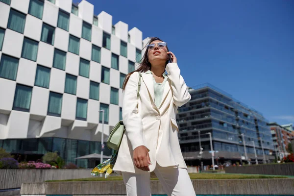 Attractive businesswoman in white suit walking on skyscrapers cityscape background