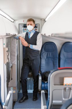 Full length of man airline worker in protective face mask standing near passenger seat in aircraft salon