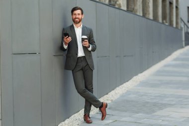 Businessman in suit with phone drinking coffee near office building. Business concept