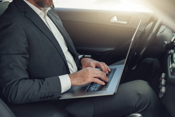 Close up of businessman working on laptop while sitting on driver seat in car. Blurred background