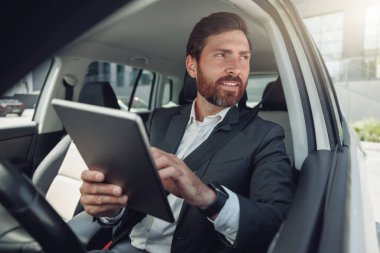 Businessman working on digital tablet while sitting on driver seat in car. Blurred background