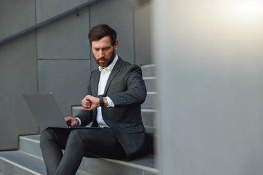 Businessman in suit working laptop and looking on wriste watch near office. Business concept
