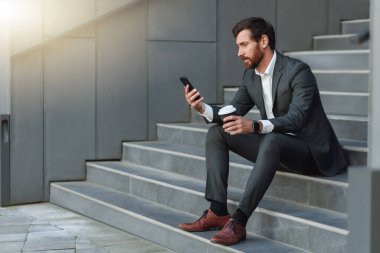 Businessman in suit with phone drinking coffee near office building. Business concept