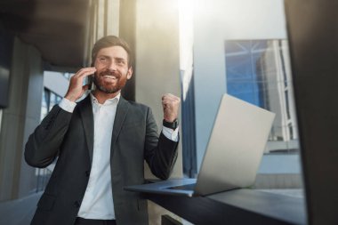 Smiling businessman in suit with laptop talking phone near office building. Business concept