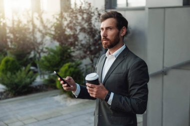 Businessman in suit with phone drinking coffee near office building. Business concept