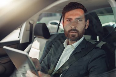 Businessman working on digital tablet while sitting on driver seat in car. Blurred background