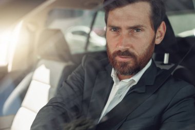 Handsome businessman in grey suit is riding behind steering wheel of car. Blurred background