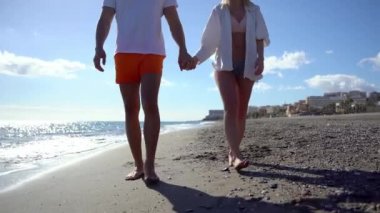Kaygısız kadın ve erkek arkadaşı gün batımında kumlu sahilde yürüyorlar ve el ele tutuşuyorlar.
