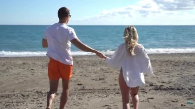 Happy couple of male and lady wearing white shirts running along sandy beach on the residing waves and holding hands