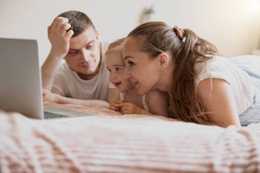 Mum dad and little daughter having fun together watching cartoons on laptop lie on sofa at home