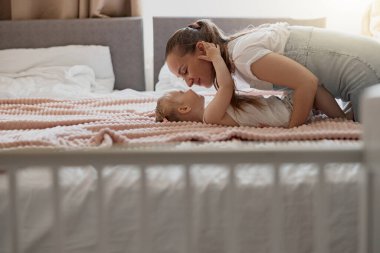 Young mother smiling and kiss her baby while lying in bed at home. Blurred background