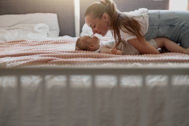 Young mother smiling and kiss her baby while lying in bed at home. Blurred background