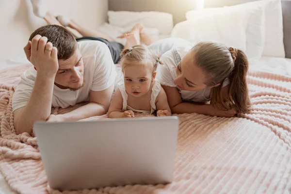 Mum dad and little daughter having fun together watching cartoons on laptop lie on sofa at home