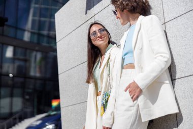 Two woman friends have a rest after walking in city standing near wall and looking each other
