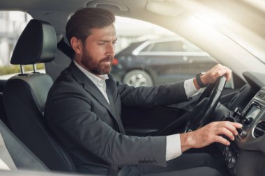 Handsome businessman in grey suit is riding behind steering wheel of car. Blurred background
