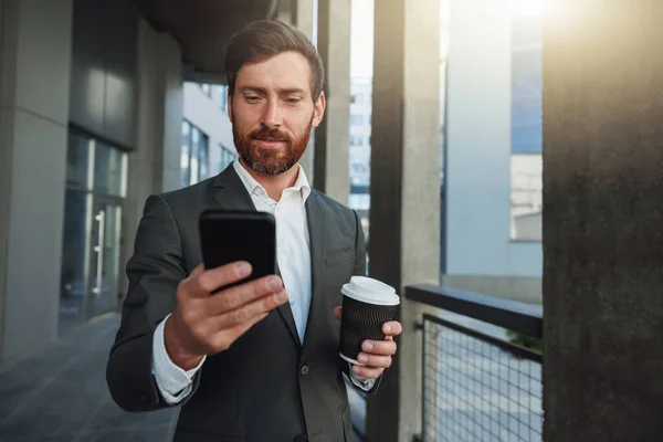 Attractive businessman holding phone standing near office building during break. Business concept