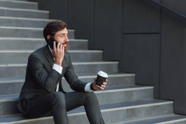 Businessman in suit sitting on stairs with coffee during break and talking phone