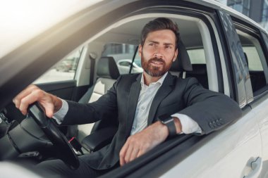 Handsome businessman in grey suit is riding behind steering wheel of car. Blurred background