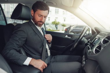 Handsome businessman is fastening his seat belt sitting in car. Blurred background