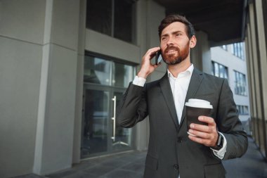 Smiling businessman in suit with coffee talking phone near office building. Business concept