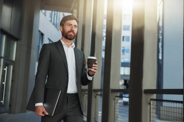 Attractive businessman with coffee and laptop near office building during break. Blurred background