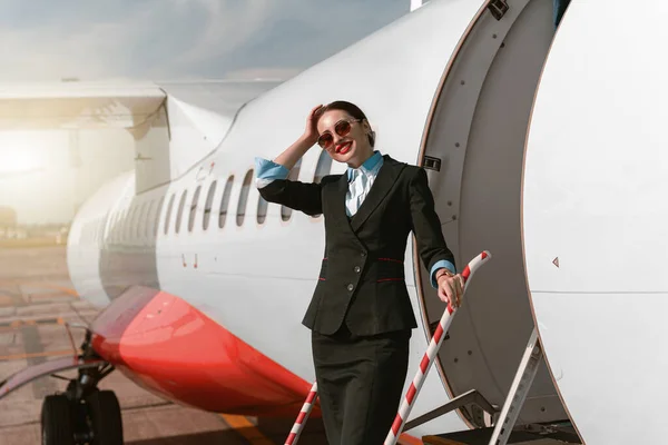 Woman flight attendant in sunglasses standing on airplane stairs at airport. Blurred background