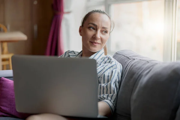 Female freelancer in casual clothes sitting on sofa and using laptop at home. Blurred background