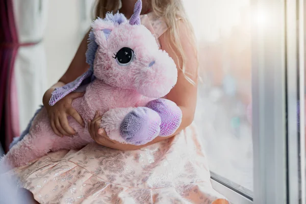 Little girl holding her soft toy unicorn while sit near window. Blurred background