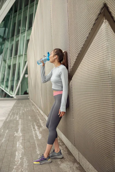 stock image Fitness woman drinking water from a bottle after workout leaning on the wall. High quality photo