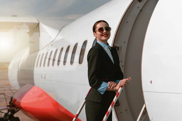 Woman flight attendant in sunglasses standing on airplane stairs at airport. Blurred background