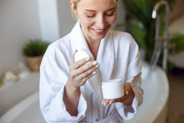 Close up of joyful blonde woman in gown opening cream or body lotion after bath taking. Beauty care.