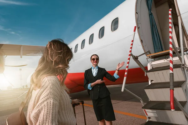 Woman stewardess standing near airplane and inviting on board. Blurred background