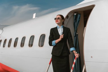Woman flight attendant in sunglasses standing on airplane stairs at airport. Blurred background