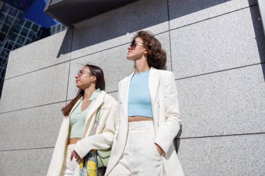 İki kadın arkadaş şehirde yürüdükten sonra dinleniyorlar. Duvarın yanında duruyorlar ve gözlerini kaçırıyorlar.