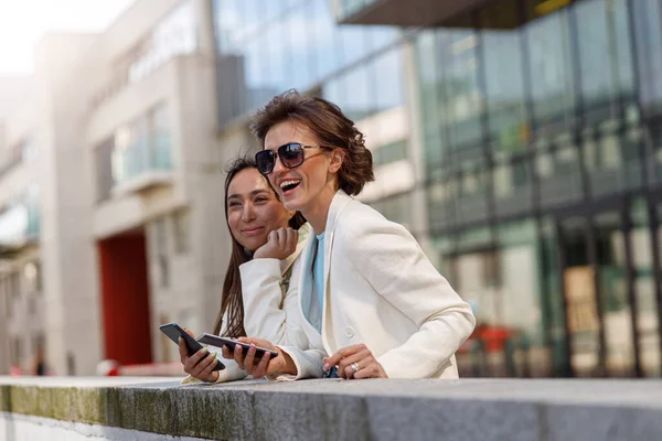 Smiling female friends in trendy outfits use their phones while relaxing in city. Blurred background