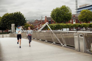 Two women in sportswear is jogging on a bridge and listening music. Running training in the city