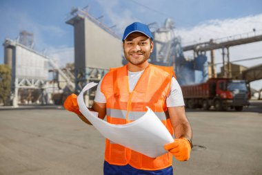 Koruyucu eldiven ve yelek giyen mutlu endüstri mühendisi, inşaat fabrikasının önünde elinde planla duruyor.