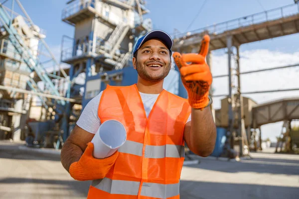 Happy multiethnic engineer wearing reflective vest and gloves, holding rolled-up blueprint while standing in front of construction material factory