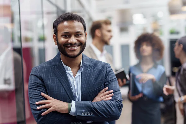 Handsome indian businessman standing in modern office on colleagues background and looking camera