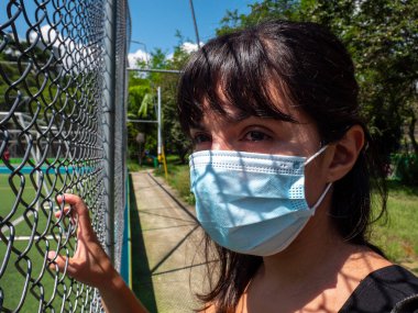 Young Brunette Woman Using a Blue Mask is Looking Through the Mesh of the Soccer Field