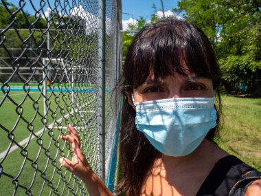 Young Brunette Woman Using a Blue Mask is Looking at the Camera and Holding the Mesh of the Soccer Field