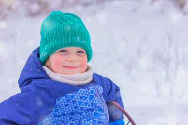 Karlı bir sokakta yeşil örgü şapkalı gülümseyen bir çocuğun portresi. Kış tatili, açık hava aktiviteleri karda, çocuklukta.