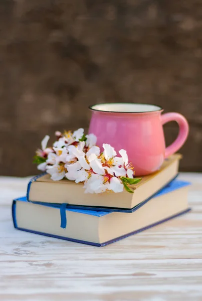 Badem ağacının çiçek açan bahar dalı ve masanın üzerindeki kitap yığınının üzerindeki pembe bardak.