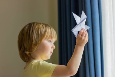 Sarı saçlı çocuk pencereden dışarı bakar ve elinde beyaz origami kağıt bir kuş tutar. Barış ve umudun sembolü, çocukluk