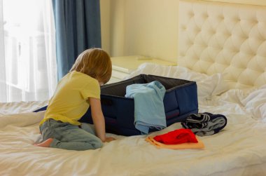 Erkek çocuk yatak odasında yatakta oturur ve açık bavula ve kıyafetlere bakar. seyahat hazırlığı, çocukluk.