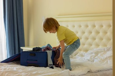 Şirin, sarışın bir çocuk giysilerini açık bir bavulun içinde yatağın üzerine katlıyor. seyahat hazırlığı, çocukluk.