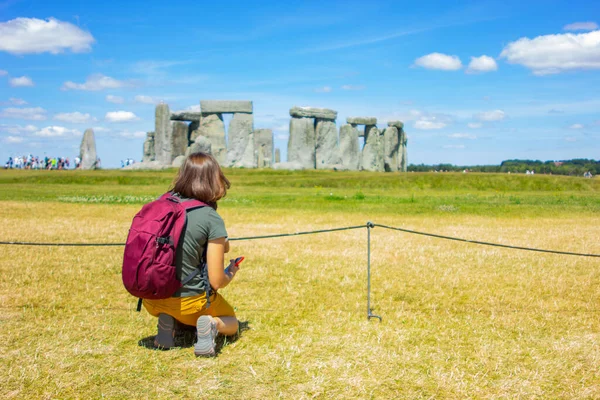 young woman tourist with a backpack sits on the background of Stonehenge, back view. England historic monument. Great Britain tourism
